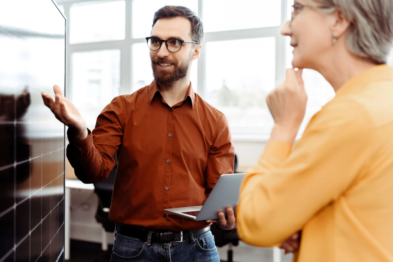 Homem de óculos escuros e camisa marrom conversando com uma mulher de cabelo grisalho e blusa amarela em ambiente de escritório, usando um tablet.