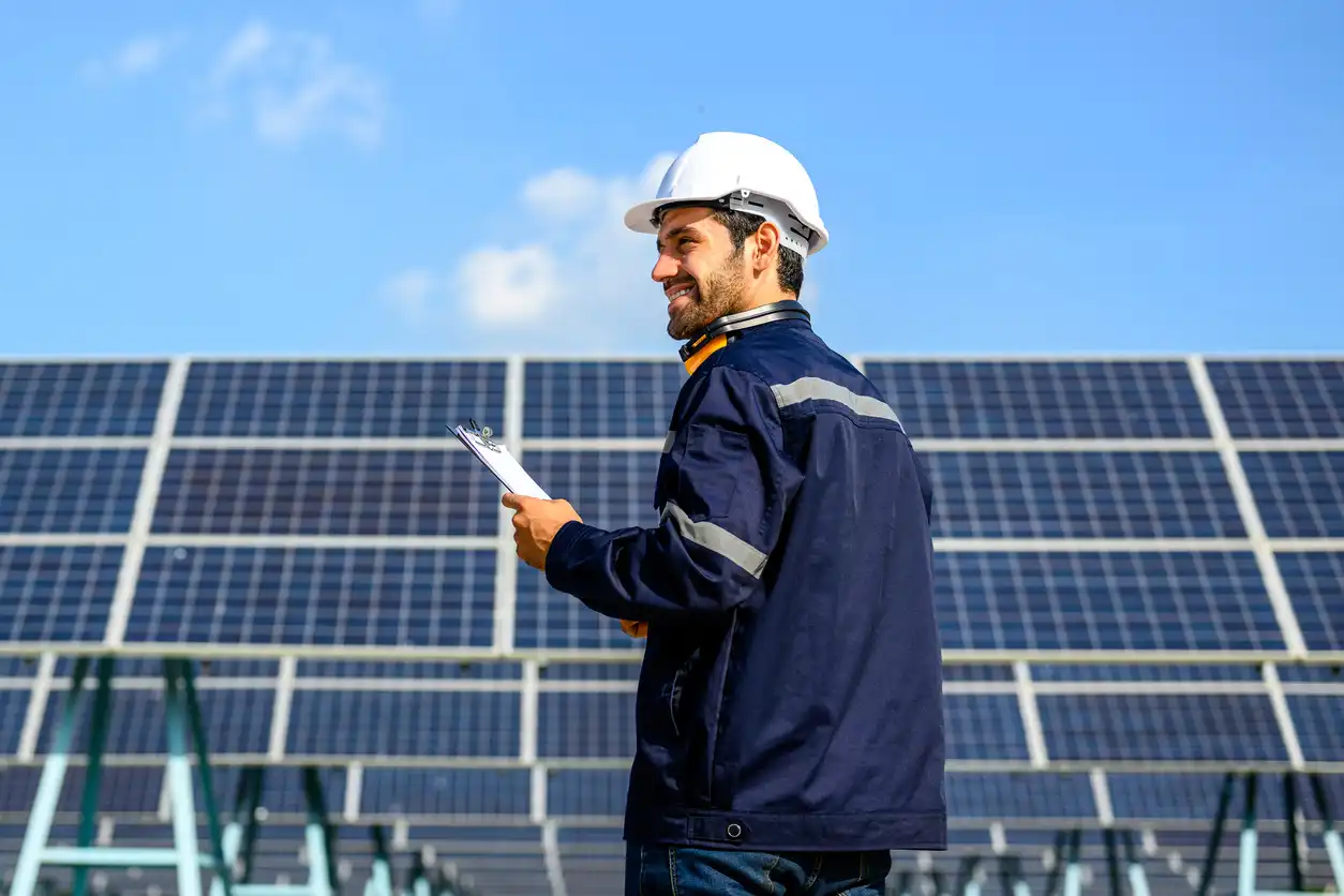 Engenheiro ou técnico de energia solar em campo, verificando painéis solares com celular, ao ar livre sob céu azul claro. Protuberância de tecnologia sustentável e energias renováveis.